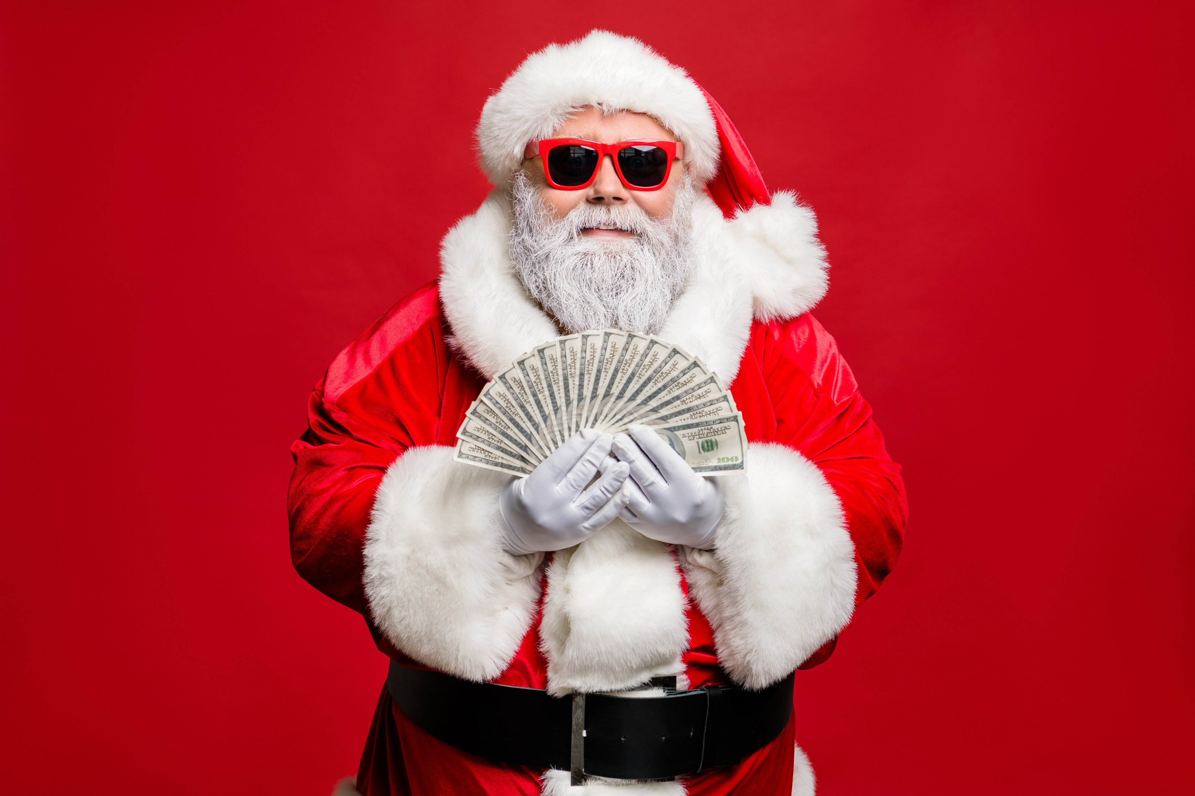 Santa Claus wearing red sunglasses and holding out money bills in a fan shape in his hands.  